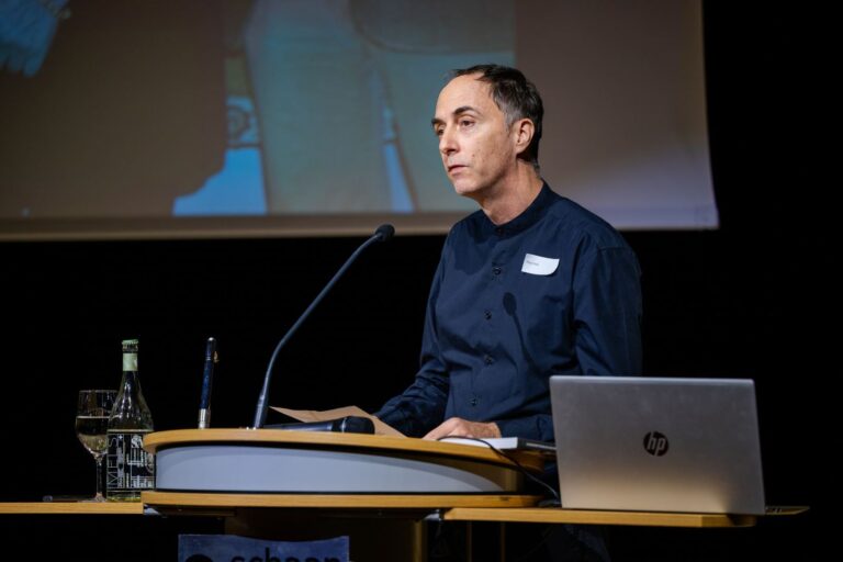 Mann spricht an einem Rednerpult mit Mikrofon, Laptop und Projektionsleinwand