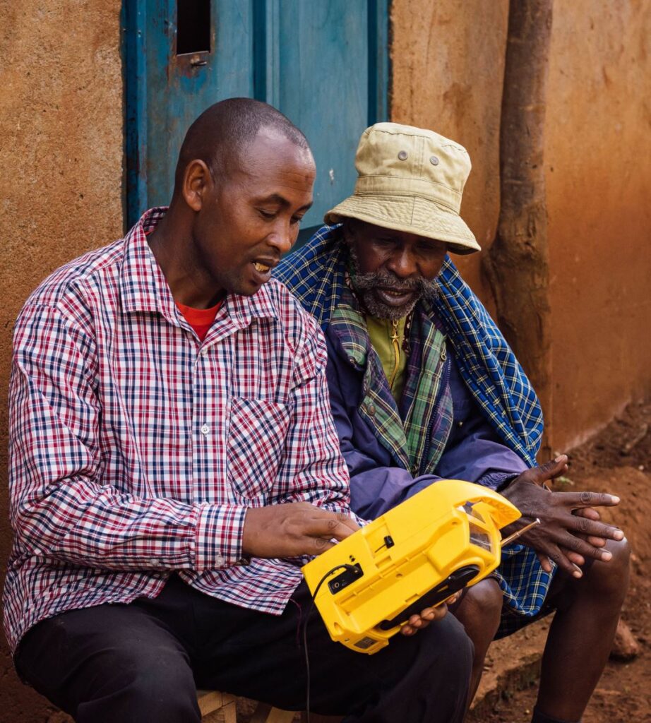 Two men discuss a yellow model vehicle outdoors beside a weathered building