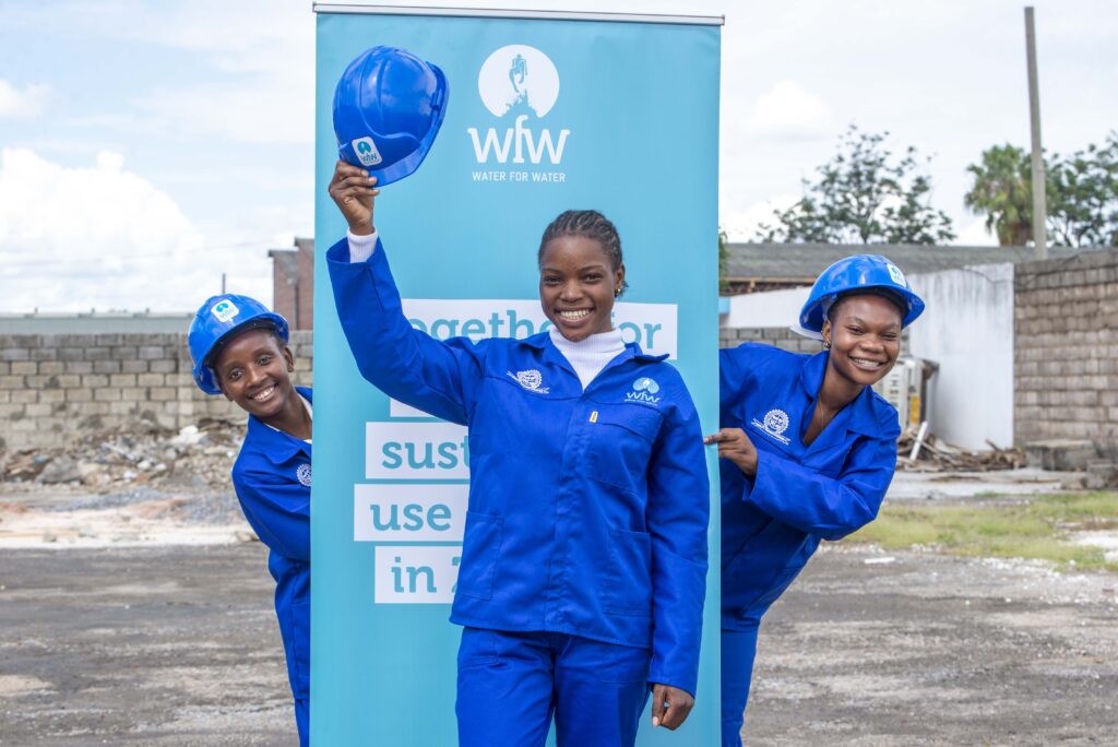 Three women in blue work uniforms and hard hats pose beside a Water for Water sign outdoors.