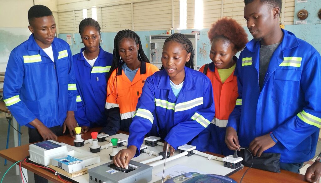 Students in work uniforms practice electrical wiring on a training board in a technical classroom