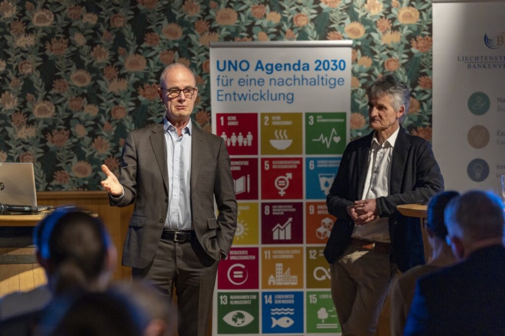 Zwei Männer präsentieren vor UNO Agenda 2030 Plakat in Konferenzraum.