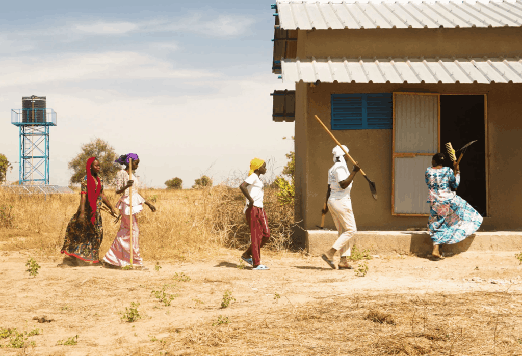 Women carrying farming tools walk toward a rural building beside a water tower