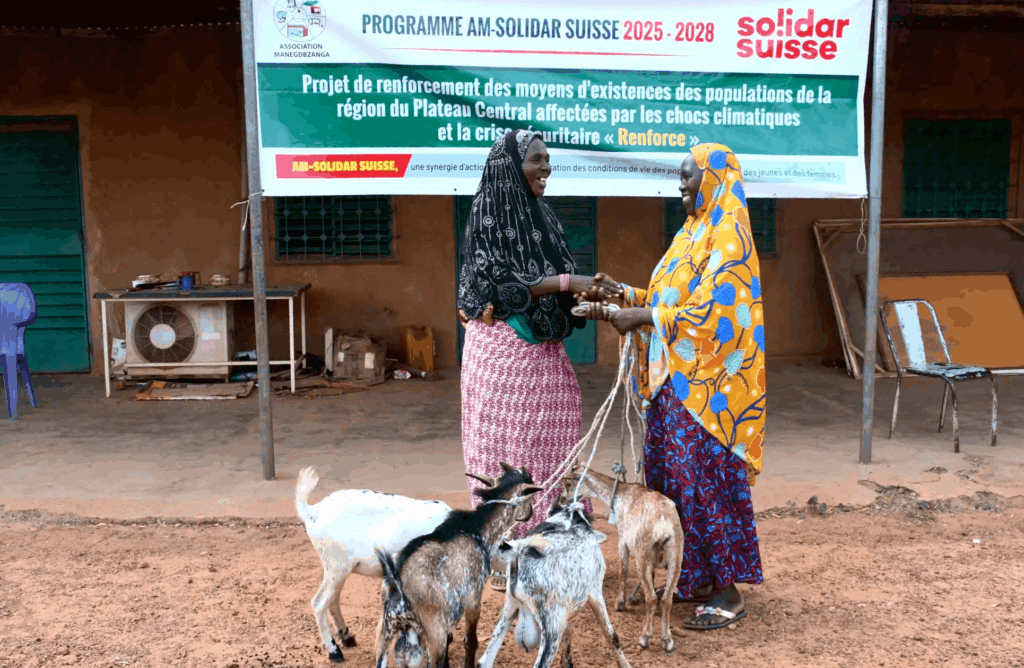 Two women shake hands beside goats outside a project banner in a rural community