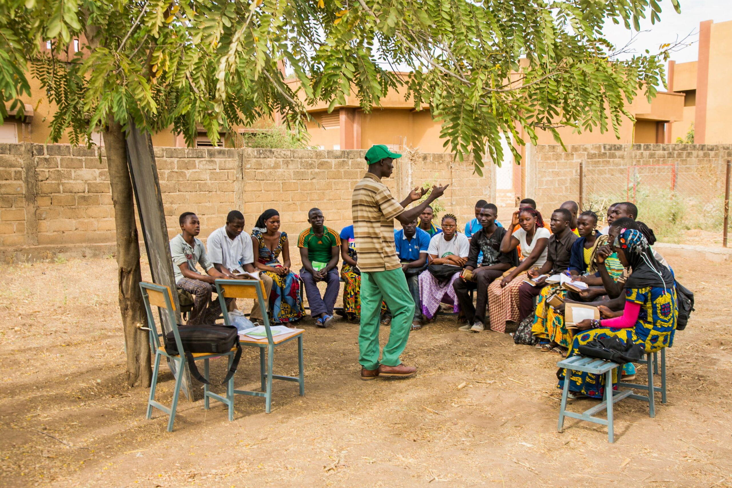 Community facilitator leads an outdoor discussion with seated youth under a tree