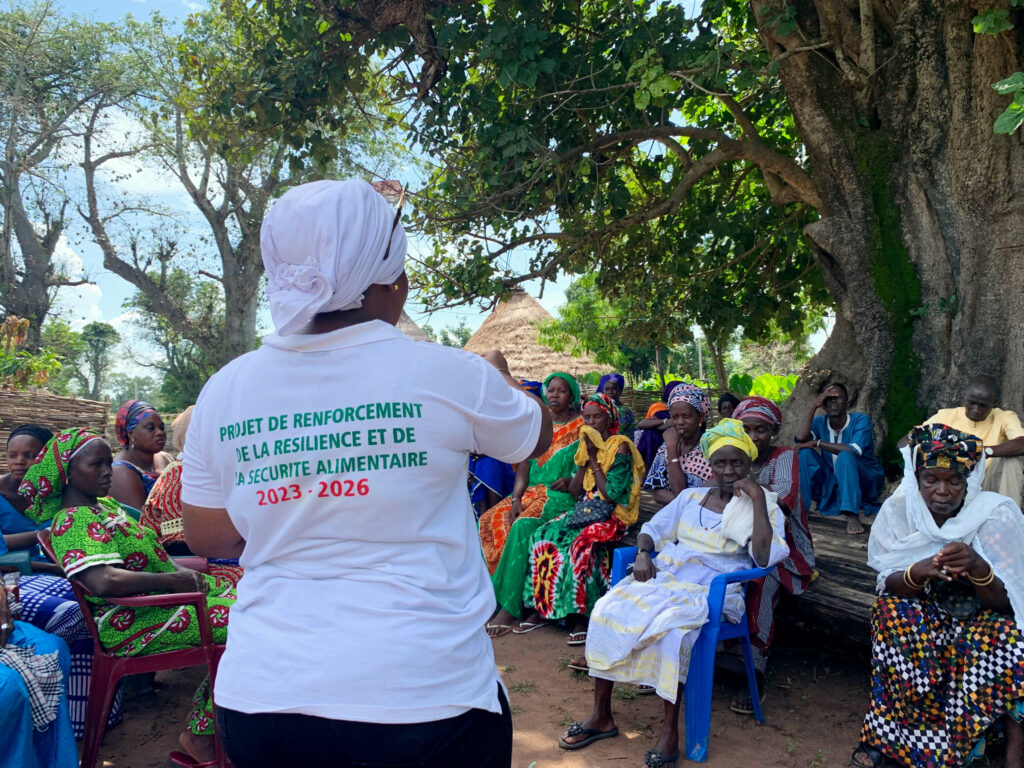 Community meeting on food security resilience project under trees, led by a woman in white