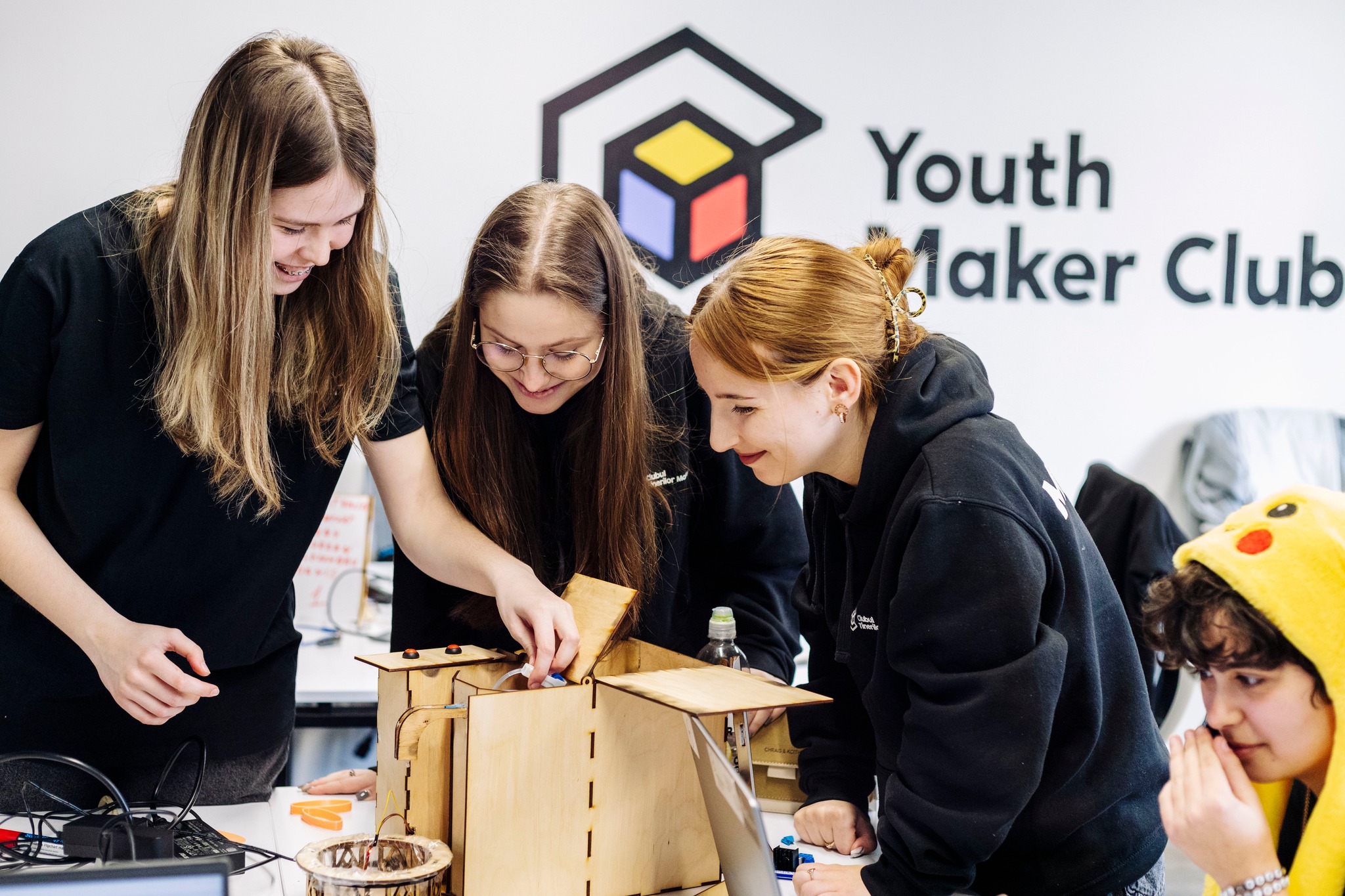 Drei junge Frauen in schwarzen Kapuzenpullis, de gemeinsam ein technisches Gerät anschauen. 