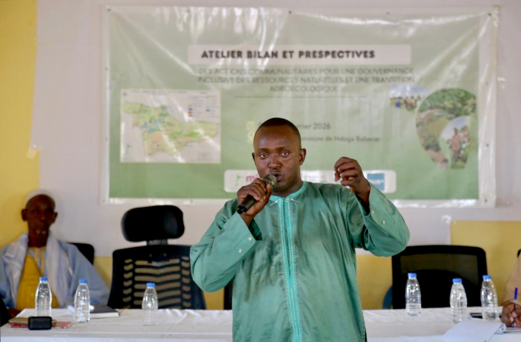 Speaker in green tunic addresses a development workshop with banner and panel table behind