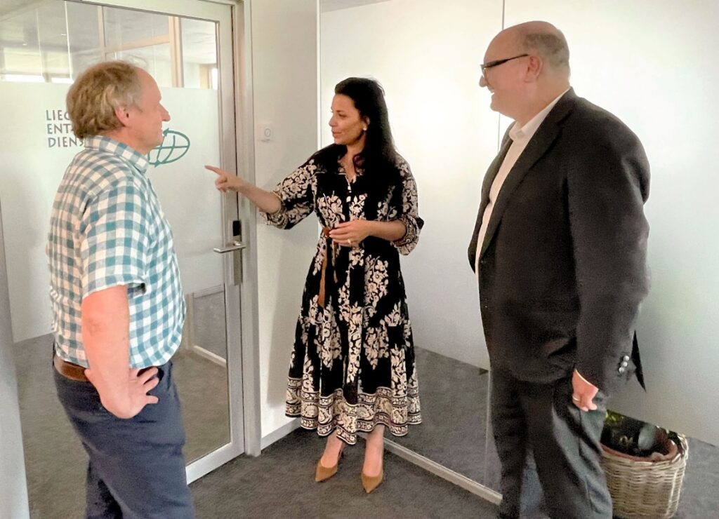 Three colleagues discuss inside LED office, with woman pointing toward a glass door