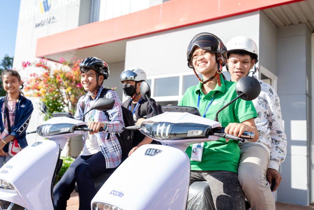 Young people wearing helmets sit on electric scooters outside a building, smiling together.