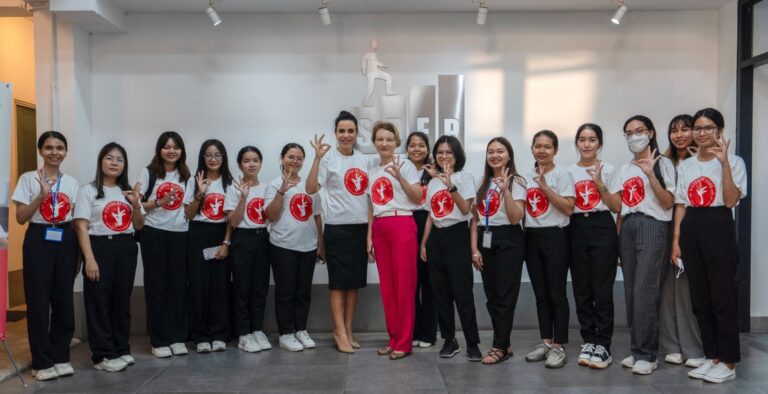 Gruppe von Frauen in weißen T-Shirts mit rotem Logo posiert lächelnd im Büro.