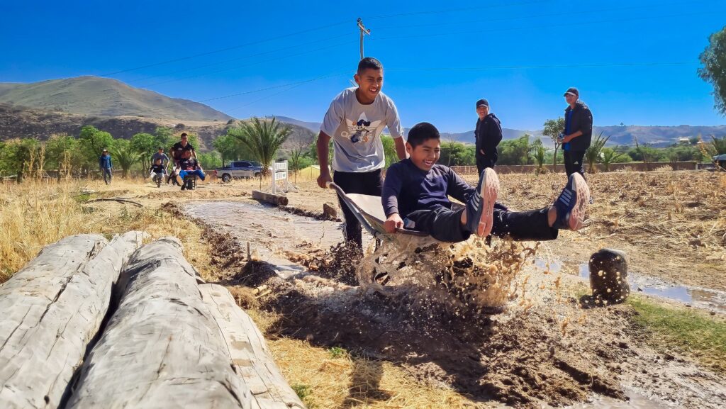 Boys push a wheelbarrow through muddy water on a rural obstacle course
