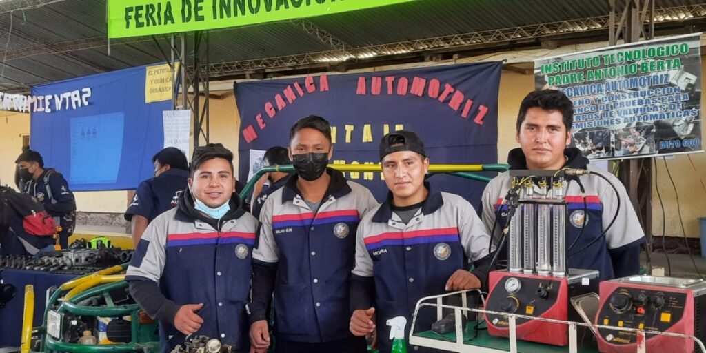 Four men in work uniforms stand beside mechanical training equipment at an innovation fair booth
