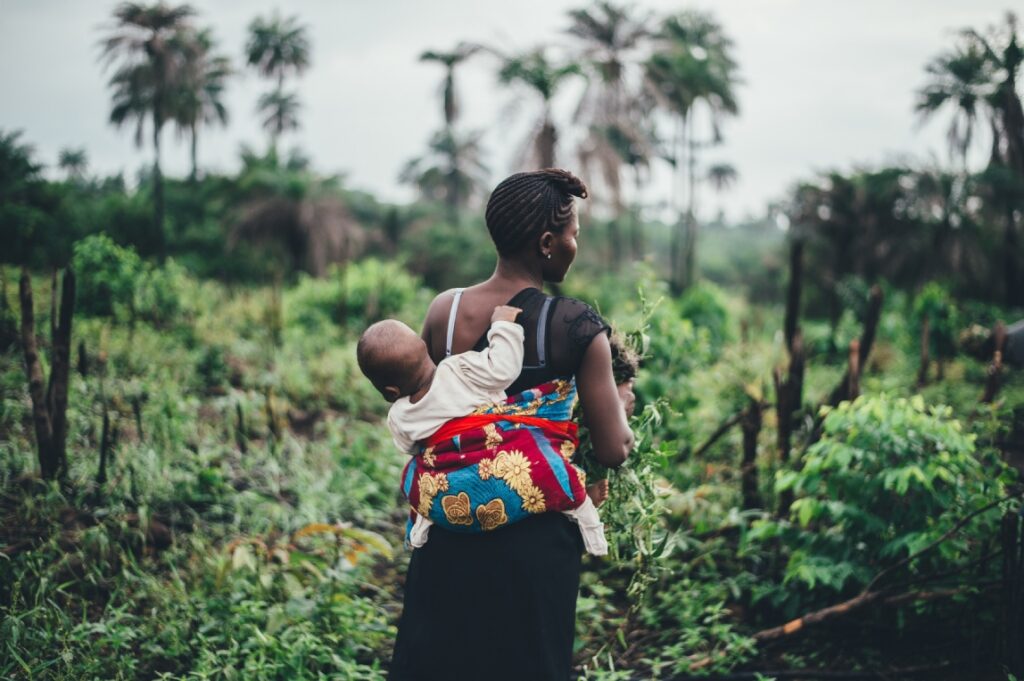 Woman carrying a child in a colorful wrap through a lush rural field with palm trees