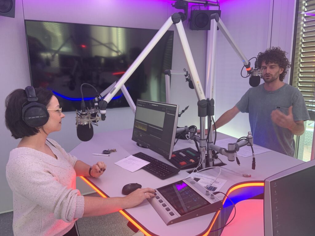 Two radio hosts record a live studio discussion with microphones, headphones, and control screens.