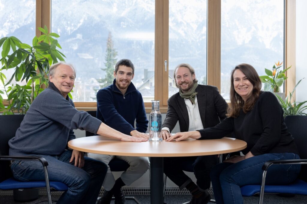 Four colleagues at a round table in an office, smiling beside a water bottle and mountain view.