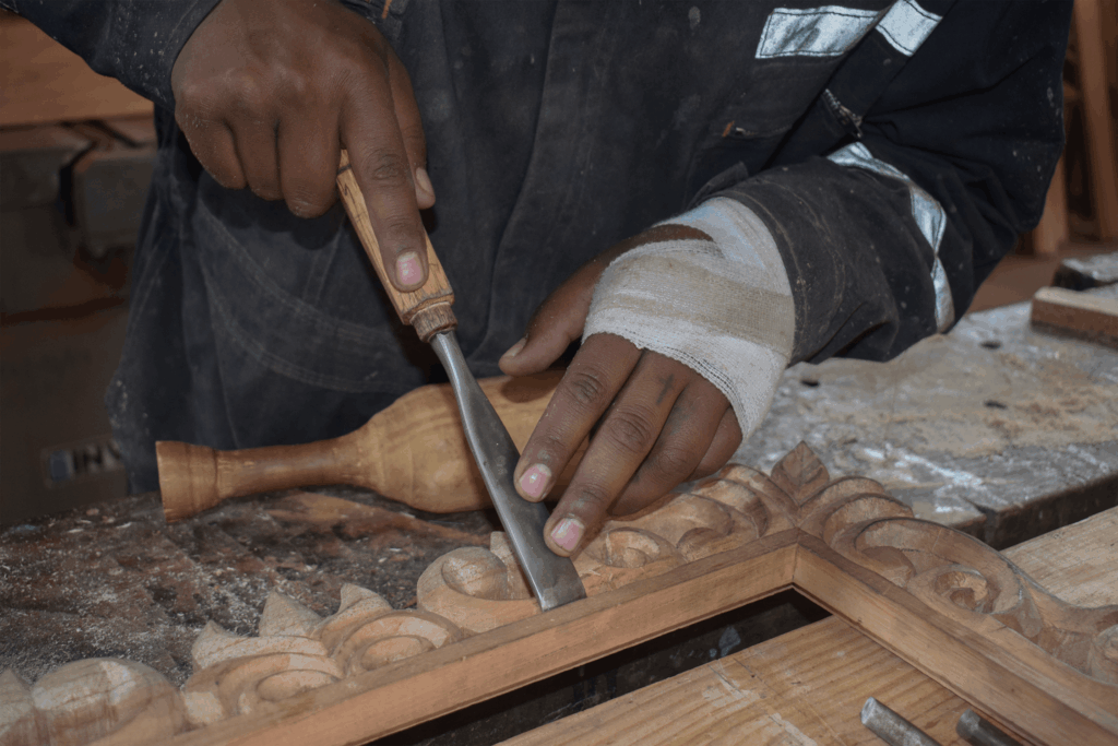Woodcarver shaping an ornate frame with a chisel, bandaged hand steadying the workpiece