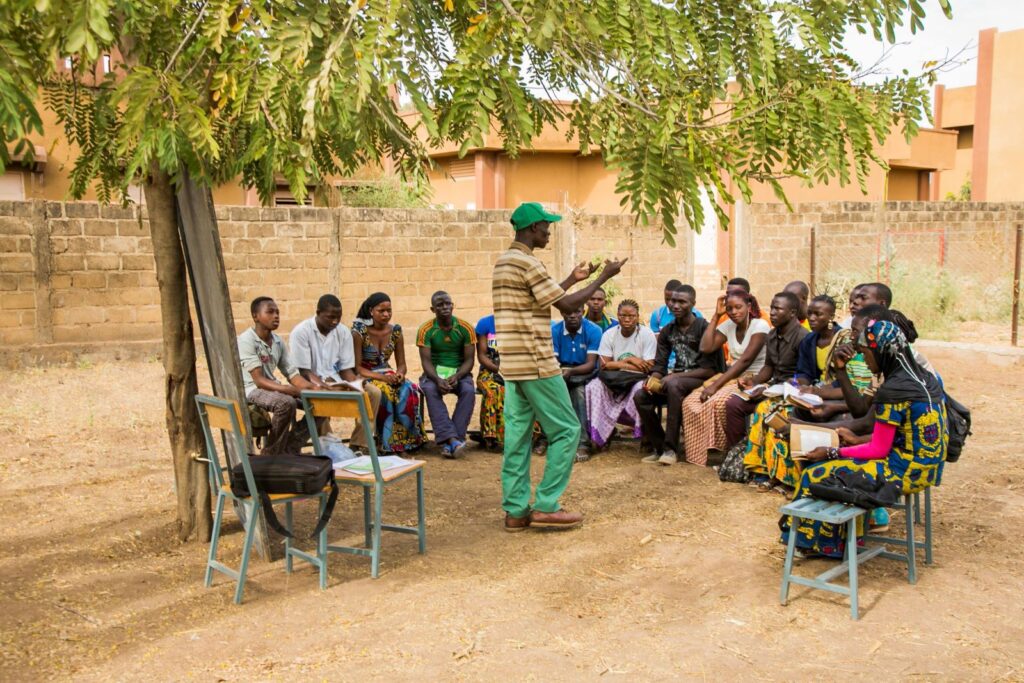 Community facilitator leads an outdoor training session with seated participants under a tree