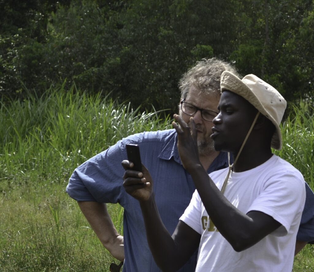 Two men discuss field observations outdoors, one holding a smartphone in tall grass