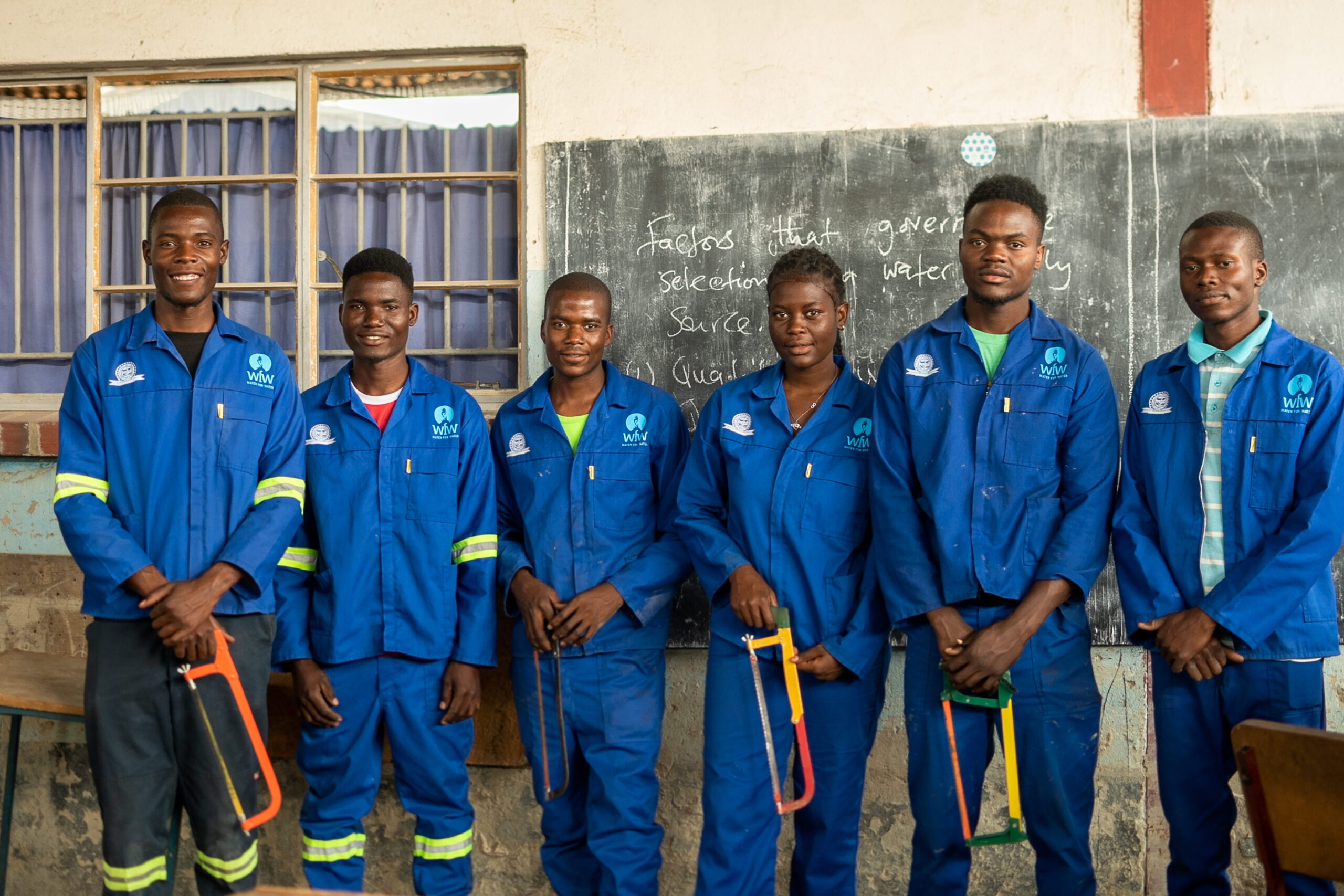 Sechs junge Erwachsene in blauen Arbeitsanzügen vor einer Tafel im Klassenzimmer