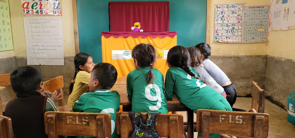Kinder sitzen in einem Klassenzimmer vor einer kleinen Puppenbühne mit LED-Logo