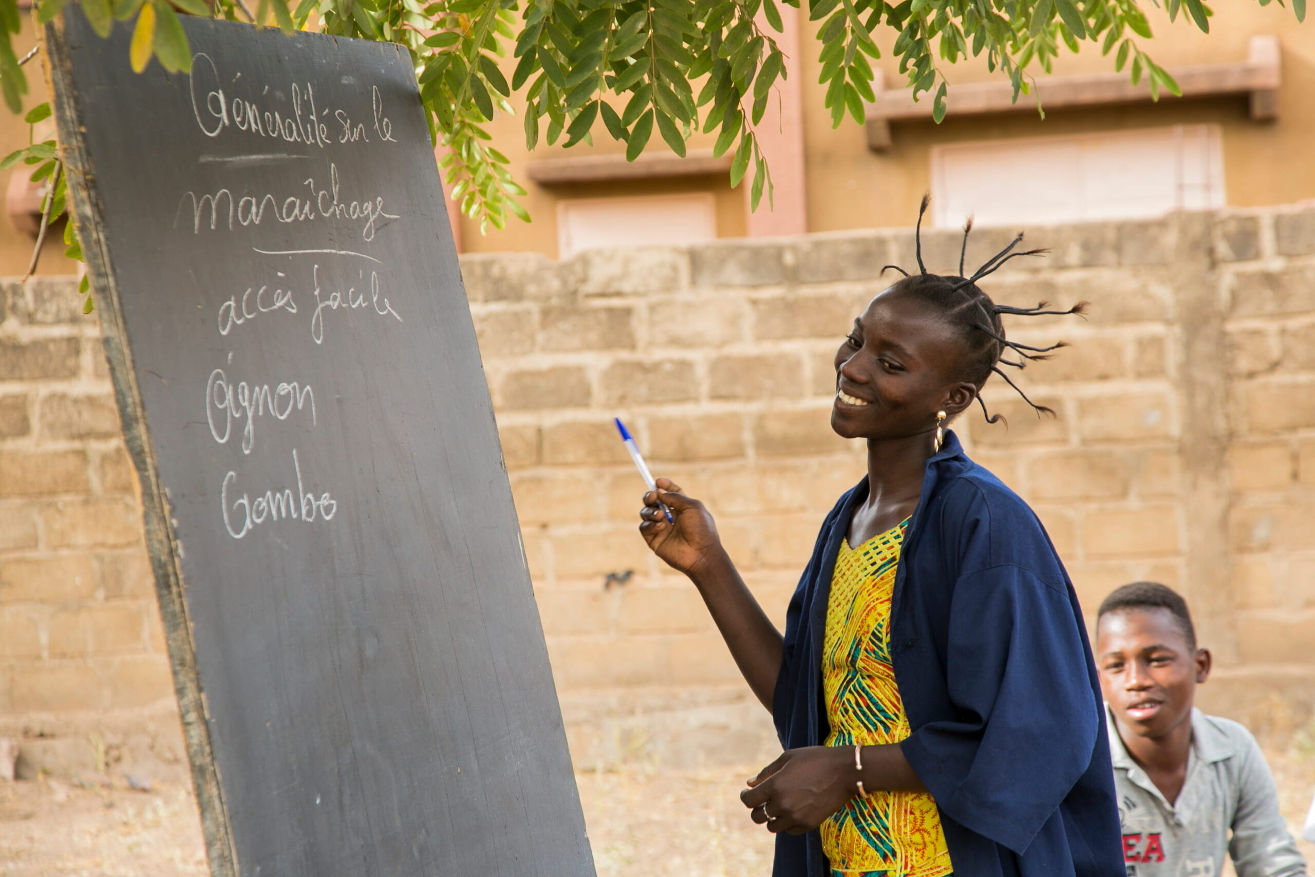Junge Frau erklärt Inhalte auf einer Tafel im Freien, Schüler im Hintergrund