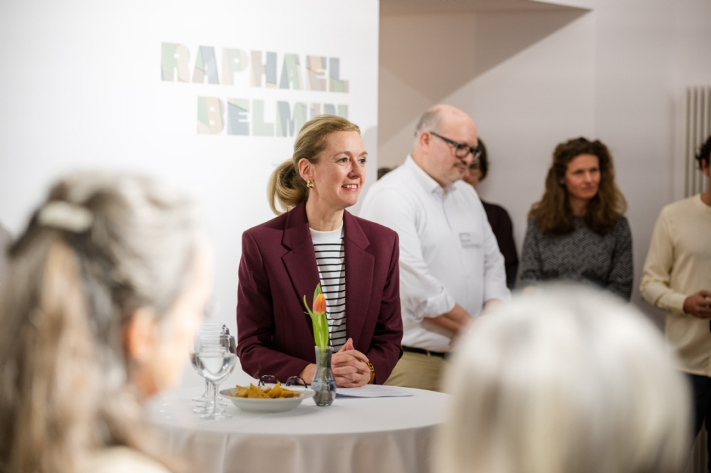 Eine Frau mit rotem Blazer und einer gestreiften Bluse, die vorne steht und zum Publikum spricht. 